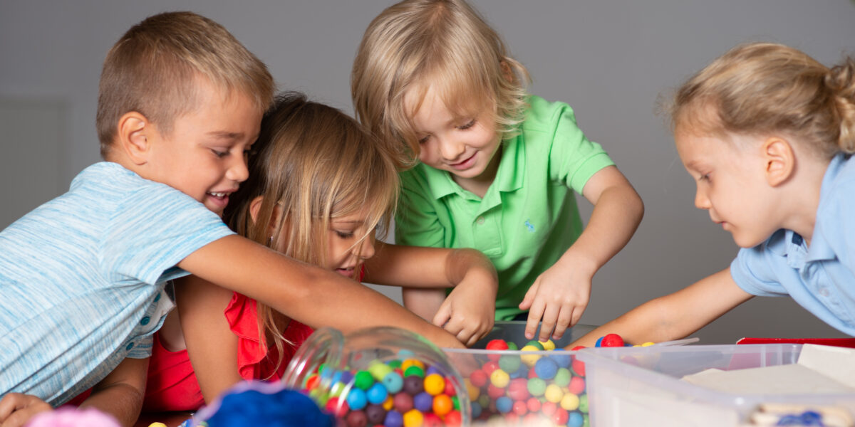 Vier kleine Kinder sind um einen Tisch versammelt und greifen in einen Behälter mit bunten Perlen oder Bonbons, wobei sie engagiert und konzentriert wirken.
