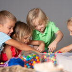 Vier kleine Kinder sind um einen Tisch versammelt und greifen in einen Behälter mit bunten Perlen oder Bonbons, wobei sie engagiert und konzentriert wirken.