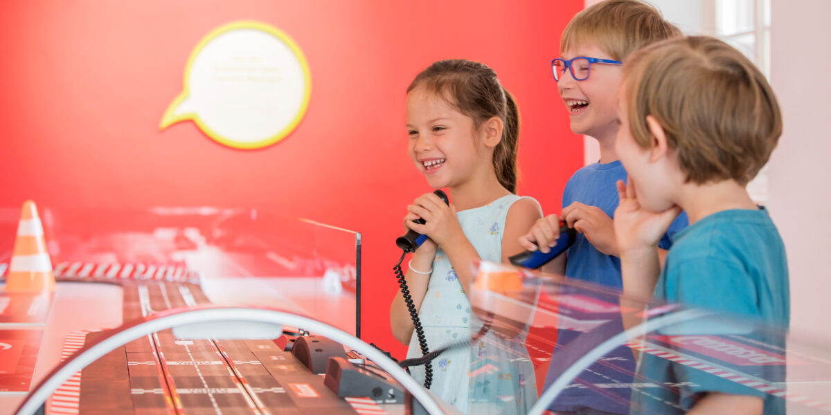 Drei kleine Kinder stehen in einem hell erleuchteten Raum mit roten Wänden um eine Slotcar-Rennbahn herum, lächeln und halten Controller in der Hand.