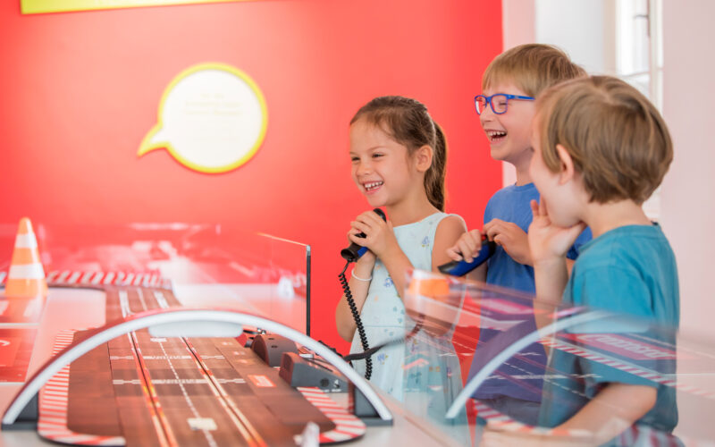 Drei kleine Kinder stehen in einem hell erleuchteten Raum mit roten Wänden um eine Slotcar-Rennbahn herum, lächeln und halten Controller in der Hand.