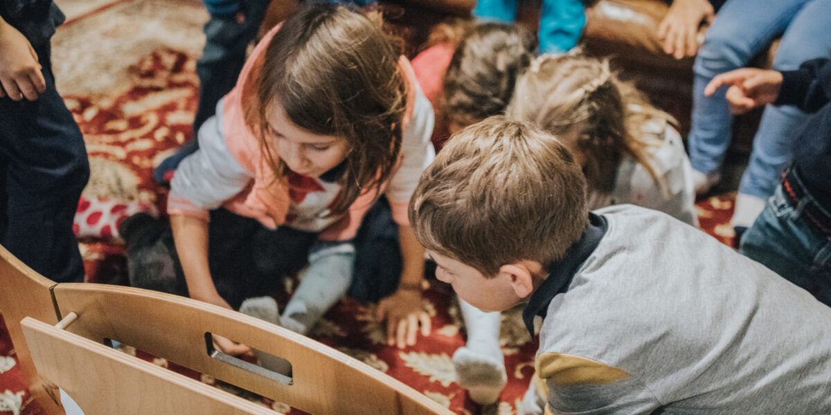 Mehrere Kinder sitzen auf einem gemusterten Teppich, dicht gedrängt um einen hölzernen Gegenstand herum und scheinen konzentriert einer Aktivität im Haus nachzugehen.