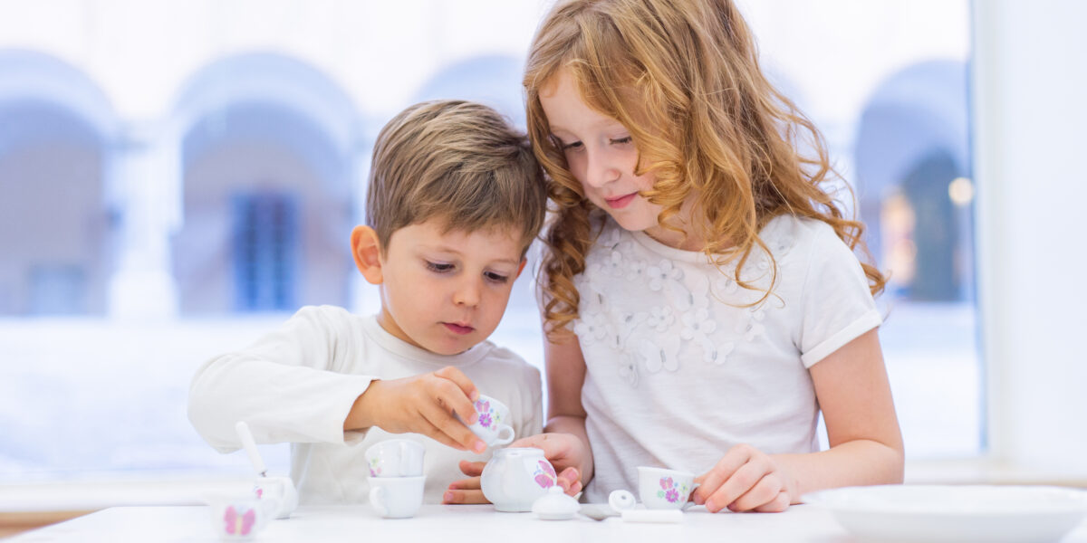 Zwei Kinder sitzen an einem Tisch und feiern eine Teeparty mit einem kleinen weißen und geblümten Spielzeug-Teeservice.