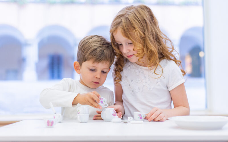 Zwei Kinder sitzen an einem Tisch und feiern eine Teeparty mit einem kleinen weißen und geblümten Spielzeug-Teeservice.