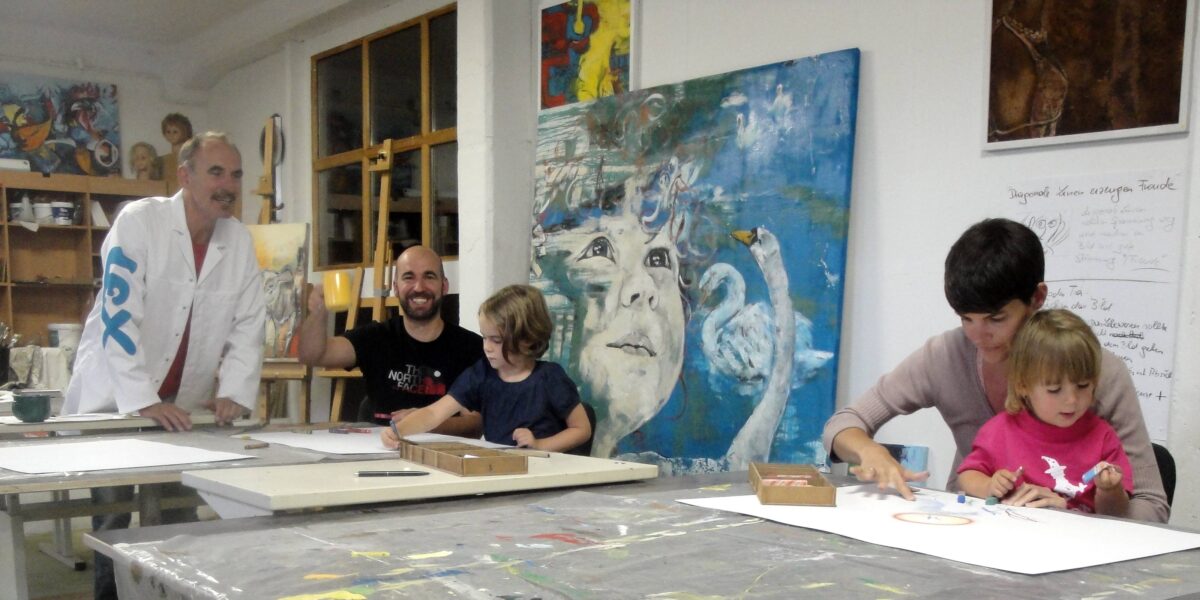 Eine Gruppe von Erwachsenen und Kindern sitzt an Tischen und zeichnet in einem Kunstatelier, umgeben von Gemälden und Kunstzubehör.