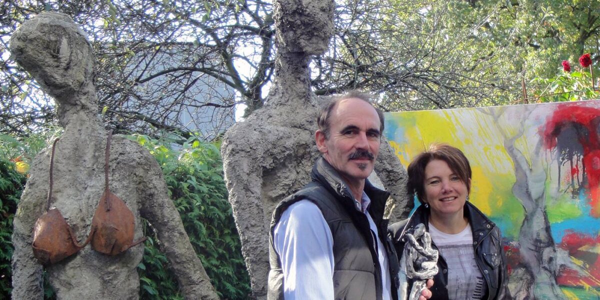 Ein Mann und eine Frau stehen im Freien neben zwei grob strukturierten menschlichen Skulpturen und einem farbenfrohen abstrakten Gemälde, mit Bäumen und Grünpflanzen im Hintergrund.
