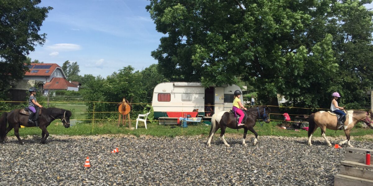 Drei Kinder reiten auf Pferden in einem eingezäunten Außenbereich mit einem Wohnwagen und Bäumen im Hintergrund. Orangefarbene Verkehrskegel markieren einen Weg auf dem Boden.