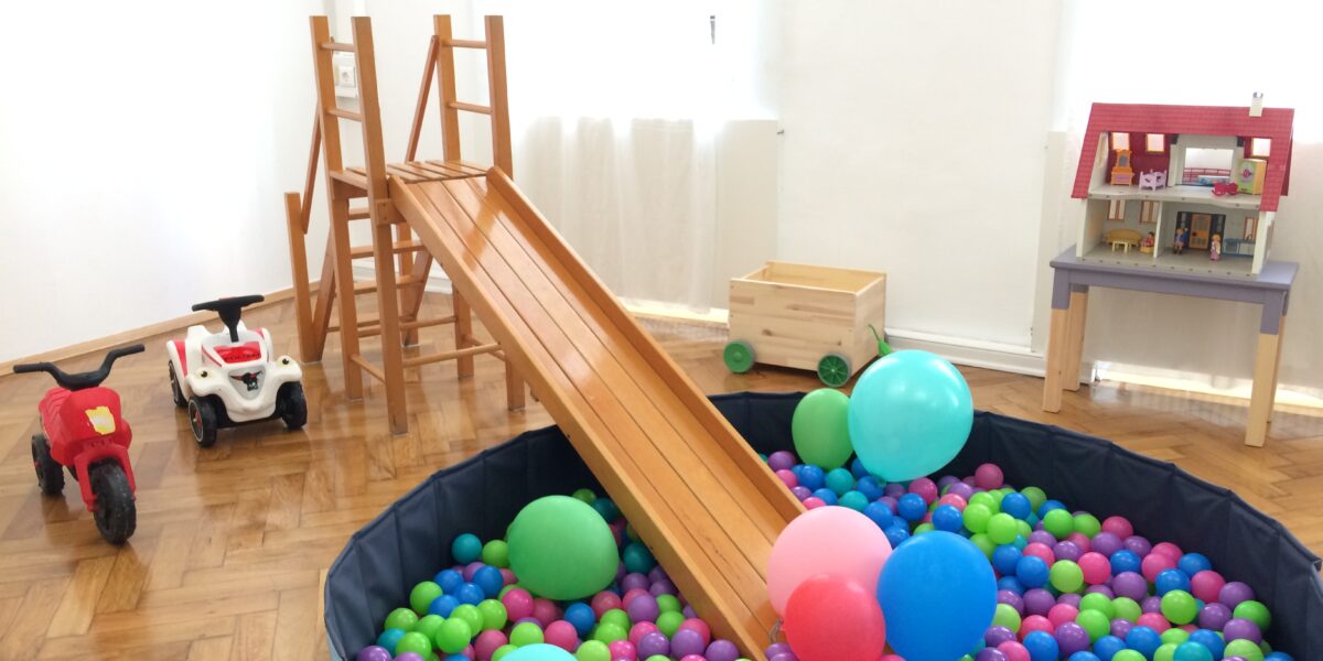 Ein Kinderspielzimmer mit einer Holzrutsche, die in ein Bällebad führt, Spielautos, Luftballons, einem Puppenhaus und einer Spielzeugkiste auf einem Holzboden.