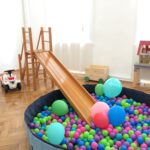 Ein Kinderspielzimmer mit einer Holzrutsche, die in ein Bällebad führt, Spielautos, Luftballons, einem Puppenhaus und einer Spielzeugkiste auf einem Holzboden.