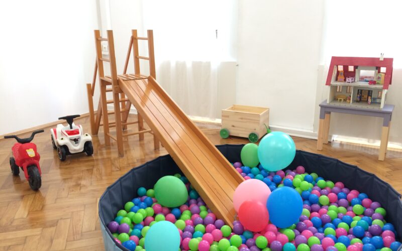 Ein Kinderspielzimmer mit einer Holzrutsche, die in ein Bällebad führt, Spielautos, Luftballons, einem Puppenhaus und einer Spielzeugkiste auf einem Holzboden.