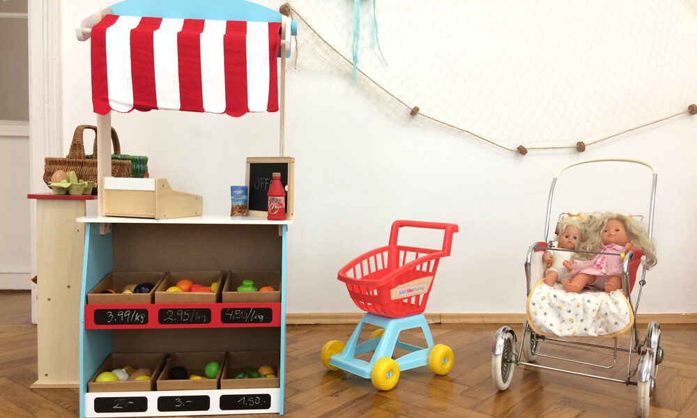 In einem Kinderspielbereich stehen vor einer weißen Wand ein Spielzeugmarktstand mit Plastikobst, ein Spielzeugeinkaufswagen und ein Puppenwagen mit zwei Puppen darin.