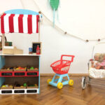 In einem Kinderspielbereich stehen vor einer weißen Wand ein Spielzeugmarktstand mit Plastikobst, ein Spielzeugeinkaufswagen und ein Puppenwagen mit zwei Puppen darin.