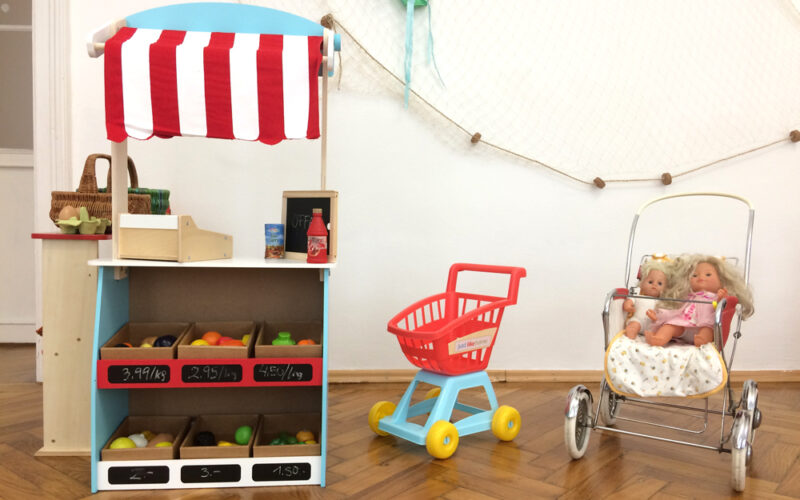 In einem Kinderspielbereich stehen vor einer weißen Wand ein Spielzeugmarktstand mit Plastikobst, ein Spielzeugeinkaufswagen und ein Puppenwagen mit zwei Puppen darin.