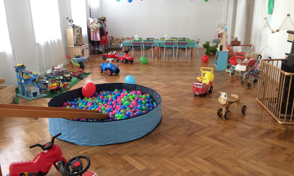 Ein Spielzimmer mit Bällebad, Spielautos und verschiedenen Kinderspielzeugen, die auf einem Holzboden verstreut sind, mit Wimpelketten dekoriert sind und im hinteren Bereich ein langer Tisch aufgestellt ist.