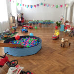 Ein Spielzimmer mit Bällebad, Spielautos und verschiedenen Kinderspielzeugen, die auf einem Holzboden verstreut sind, mit Wimpelketten dekoriert sind und im hinteren Bereich ein langer Tisch aufgestellt ist.