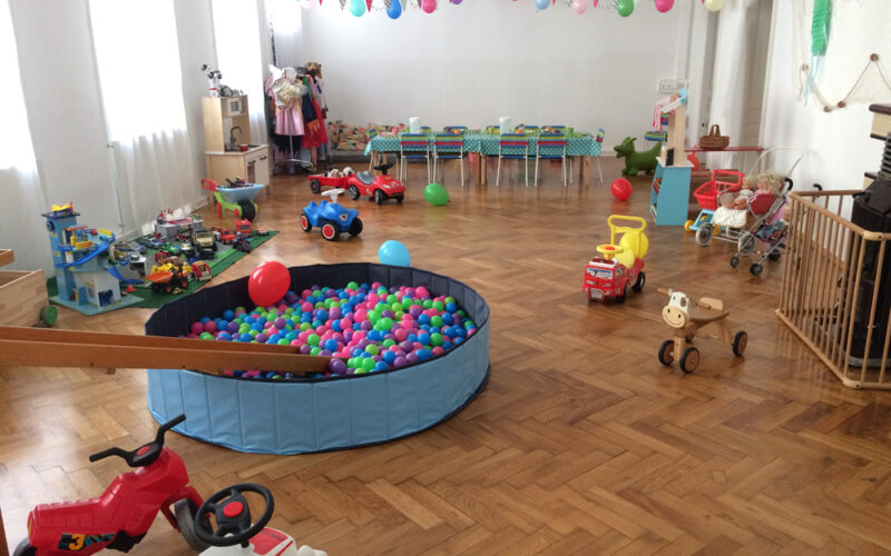Ein Spielzimmer mit Bällebad, Spielautos und verschiedenen Kinderspielzeugen, die auf einem Holzboden verstreut sind, mit Wimpelketten dekoriert sind und im hinteren Bereich ein langer Tisch aufgestellt ist.