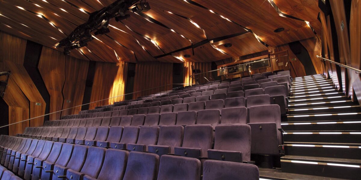 Innenansicht eines leeren Theaters mit lilafarbenen Sitzreihen, Holzwänden und -decken sowie beleuchteten Stufen im Gang.