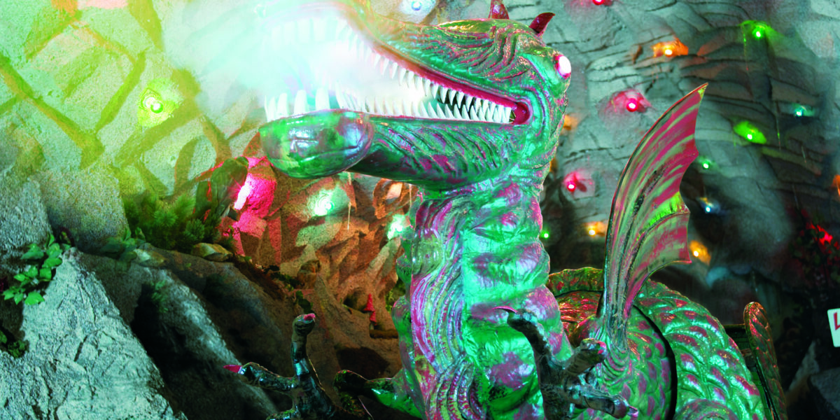 Ein großer animatronischer Drache mit leuchtenden Augen und Maul sitzt in einem Höhlentunnel, der mit bunten Lichtern dekoriert ist.