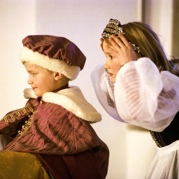 Zwei Kinder in mittelalterlichen Kostümen, von denen eines einen Spiegel hält, während das andere eine Krone auf dem Kopf zurechtrückt, spielen in einem Haus ein Rollenspiel.