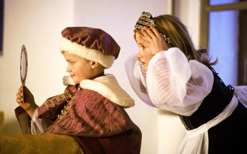 Zwei Kinder in mittelalterlichen Kostümen, von denen eines einen Spiegel hält, während das andere eine Krone auf dem Kopf zurechtrückt, spielen in einem Haus ein Rollenspiel.