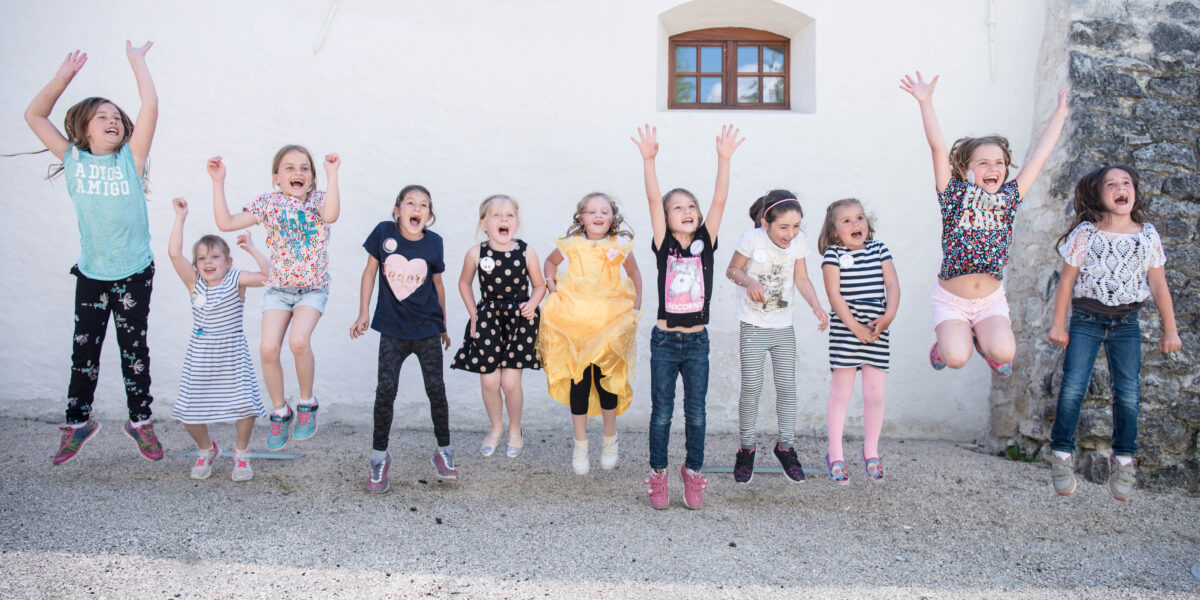 Elf kleine Kinder stehen in einer Reihe und springen draußen vor einer hellen Wand in die Luft, lächeln und spielen miteinander.