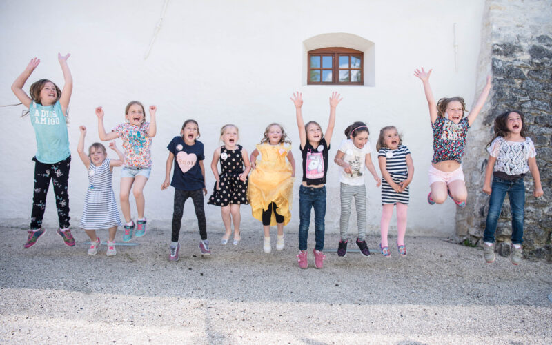 Elf kleine Kinder stehen in einer Reihe und springen draußen vor einer hellen Wand in die Luft, lächeln und spielen miteinander.