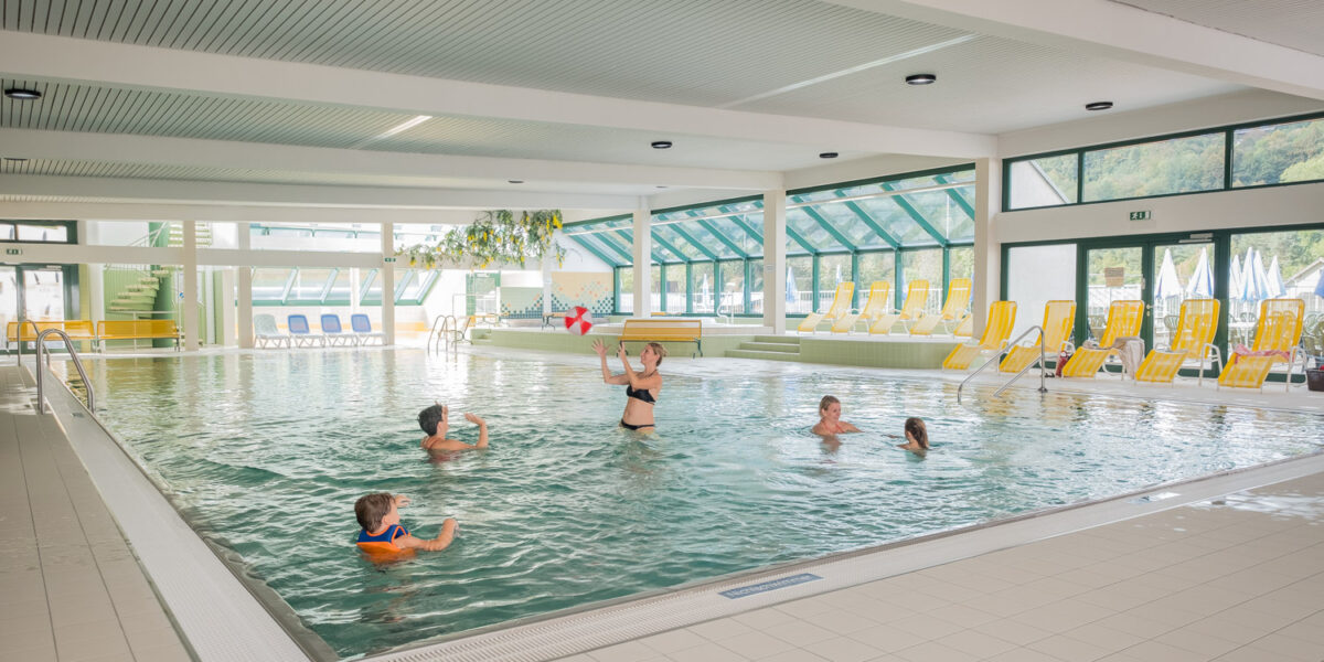 Fünf Personen schwimmen und spielen in einem großen Hallenbad mit gelben Liegestühlen an den Fenstern und am Beckenrand.