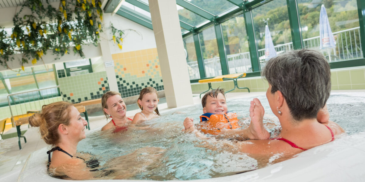 Vier Erwachsene und zwei Kinder sitzen gemeinsam in einem runden Whirlpool mit großen Fenstern, die den Blick auf das Grün und die Berge im Hintergrund freigeben.