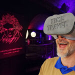 Ein Mann, der ein Virtual-Reality-Headset mit der Aufschrift "ZEITREISE" trägt, steht in einem neonbeleuchteten Raum mit einem leuchtenden Beethoven-Porträt an der Wand.