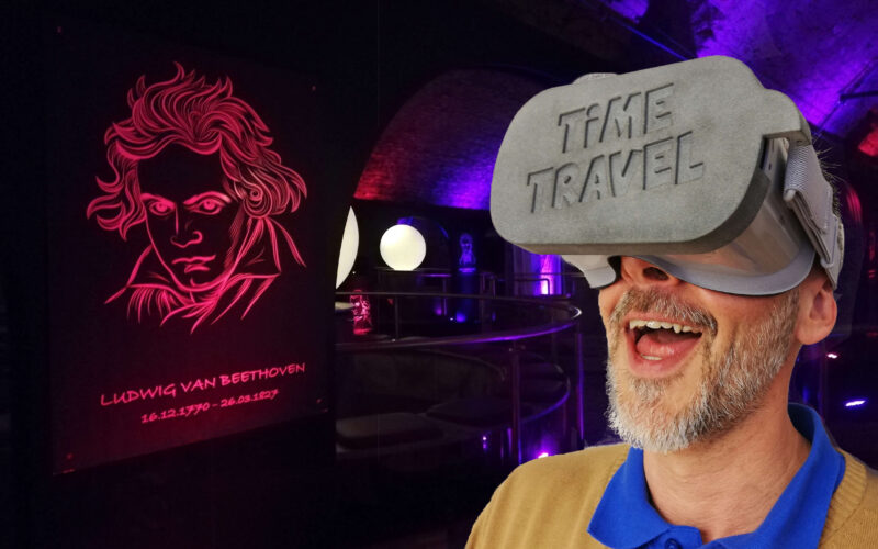 Ein Mann, der ein Virtual-Reality-Headset mit der Aufschrift "ZEITREISE" trägt, steht in einem neonbeleuchteten Raum mit einem leuchtenden Beethoven-Porträt an der Wand.