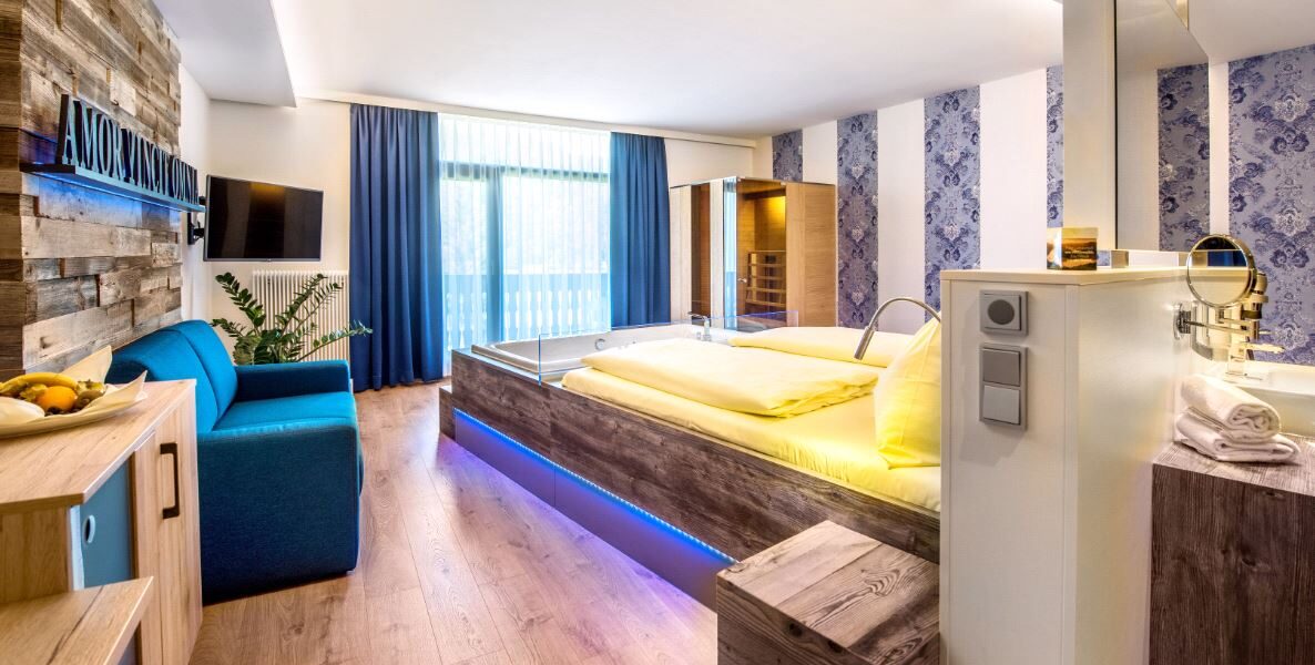 Modernes Hotelzimmer mit einem großen Bett, einem blauen Sofa, einem an der Wand montierten Fernseher, Holzfußboden und einem Fenster mit blauen Vorhängen, das Tageslicht hereinlässt.