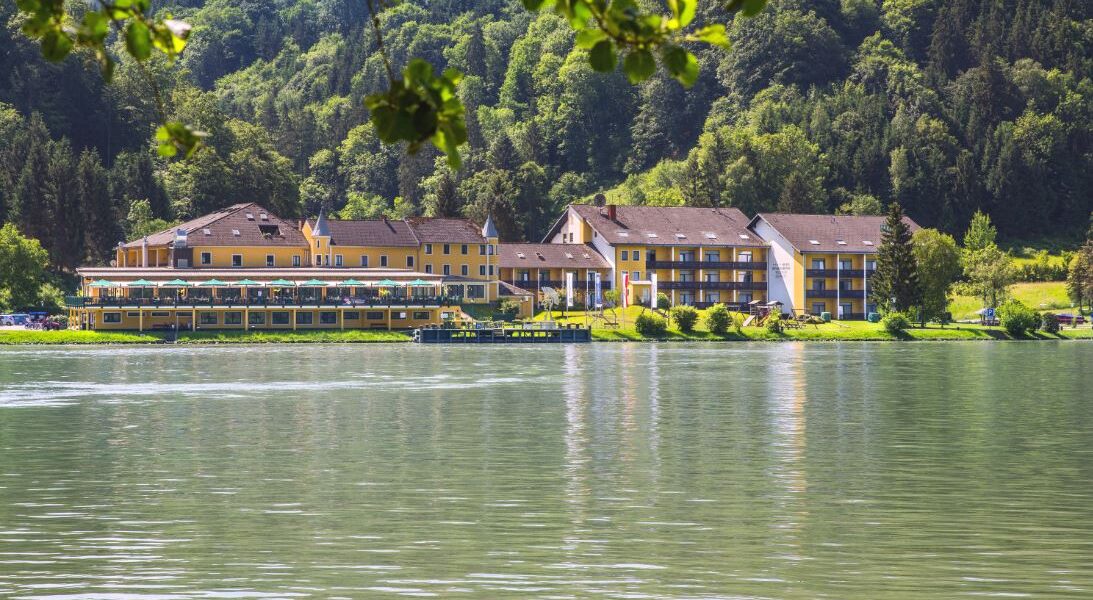 Eine Reihe gelber Gebäude, wahrscheinlich ein Hotel oder eine Ferienanlage, liegt am grünen Ufer eines ruhigen Sees mit einem bewaldeten Hügel im Hintergrund.