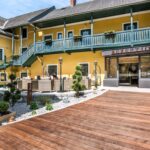 Ein gelbes, zweistöckiges Gebäude mit grünem Geländer umgibt einen Innenhof mit Sitzgelegenheiten im Freien, Topfpflanzen und einem Holzsteg, der zu einem Glaseingang mit der Aufschrift "Rezeption" führt.