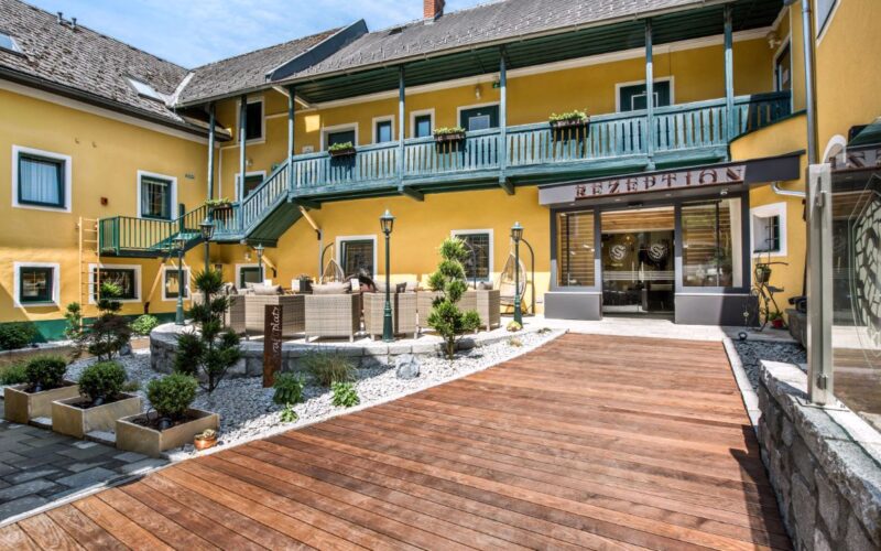 Ein gelbes, zweistöckiges Gebäude mit grünem Geländer umgibt einen Innenhof mit Sitzgelegenheiten im Freien, Topfpflanzen und einem Holzsteg, der zu einem Glaseingang mit der Aufschrift "Rezeption" führt.