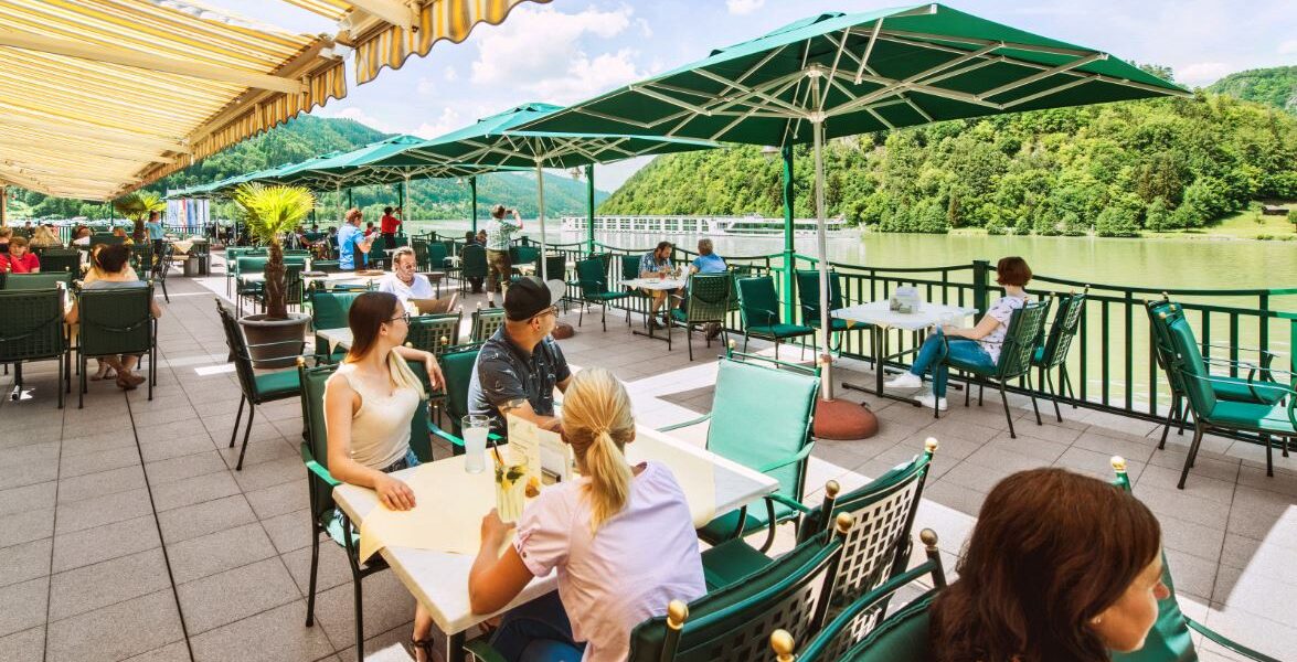 Menschen speisen im Freien unter grünen Sonnenschirmen auf einer Terrasse am Flussufer mit Blick auf die Bäume und Hügel im Hintergrund.