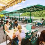 Menschen speisen im Freien unter grünen Sonnenschirmen auf einer Terrasse am Flussufer mit Blick auf die Bäume und Hügel im Hintergrund.