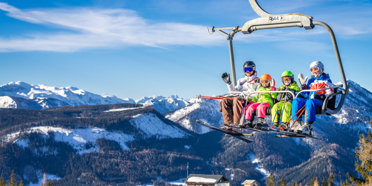 Vier Personen in bunten Skiklamotten fahren mit einem Skilift über eine verschneite Berglandschaft mit Bäumen und einem klaren blauen Himmel im Hintergrund.