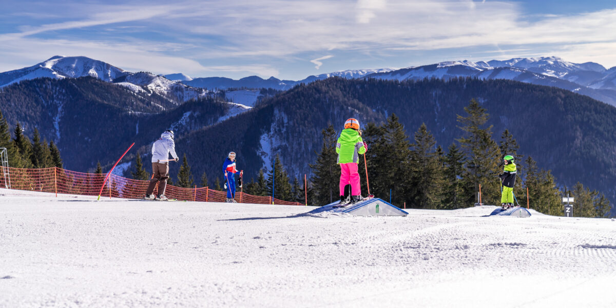 Kinder in farbenfrohen Skikostümen stehen auf einer verschneiten Piste mit Bergen und Kiefern im Hintergrund unter blauem Himmel.