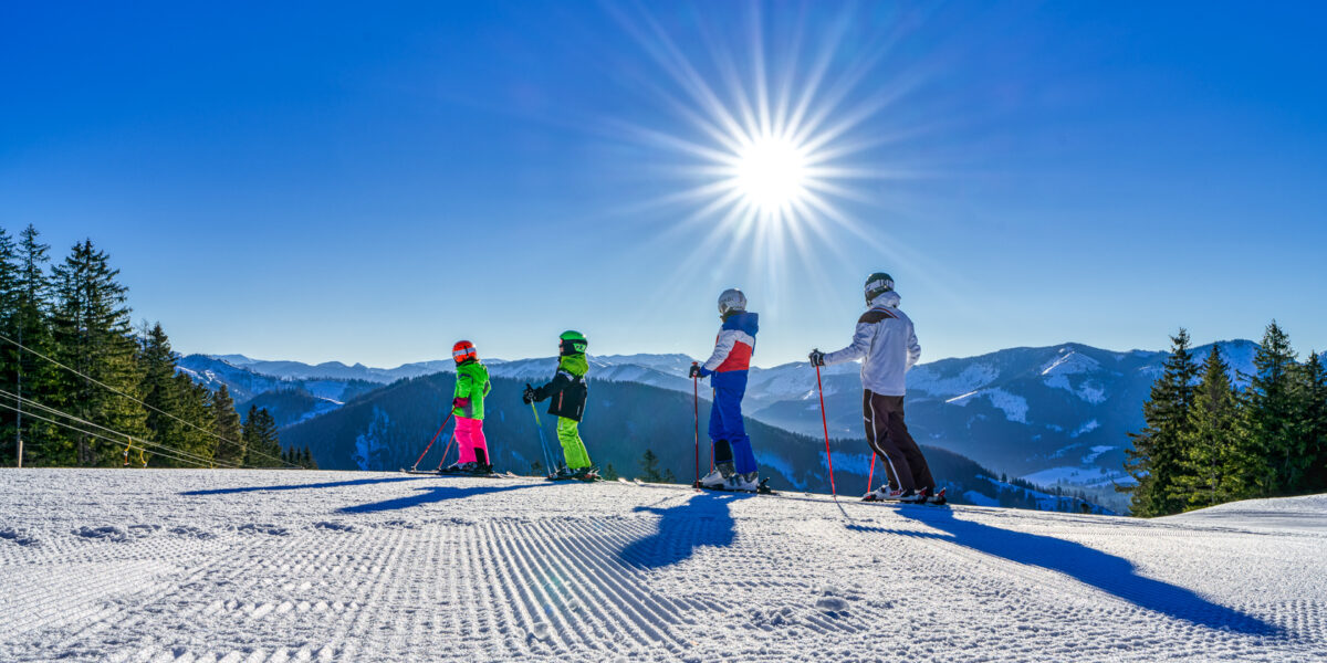 Vier Skifahrer stehen mit Skistöcken auf einer verschneiten Piste, umgeben von Bergen und Kiefern unter strahlender Sonne und blauem Himmel.
