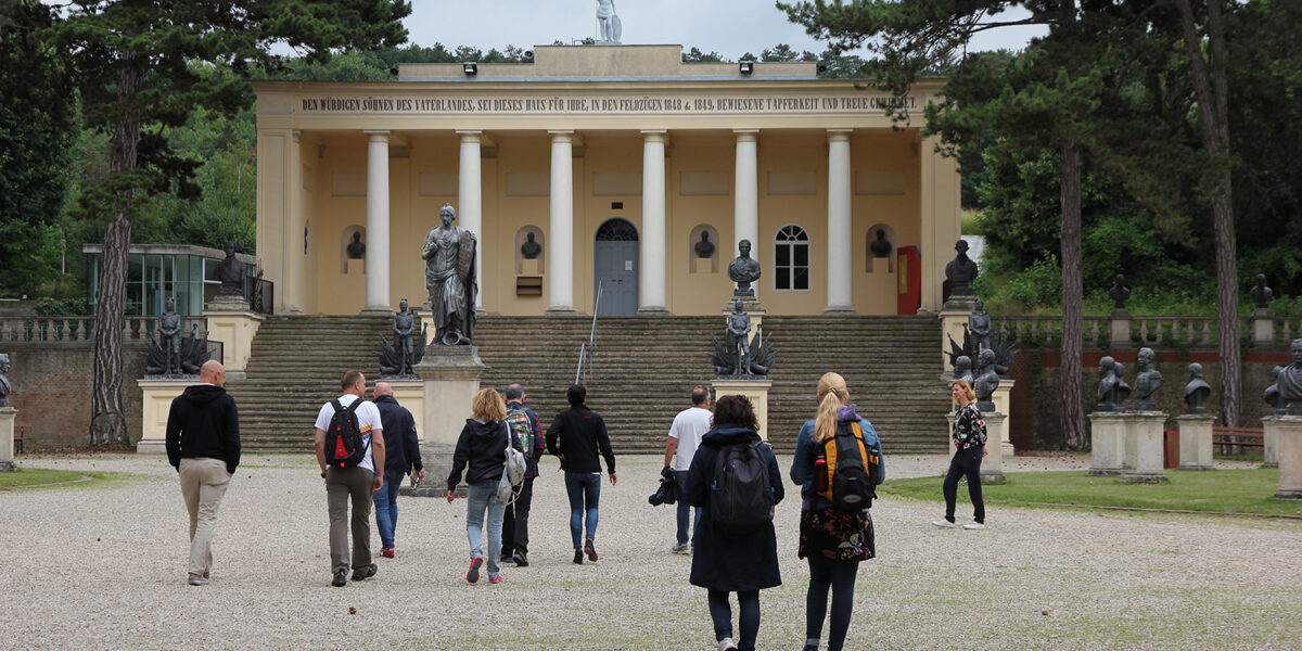 Eine Gruppe von Menschen geht auf ein neoklassisches gelbes Gebäude mit Statuen und Säulen zu, das von Bäumen und Kies umgeben ist.