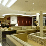 Moderne Hotellounge mit beigen Sektionssofas, Mosaikfliesenbrunnen, Ambientebeleuchtung und dekorativen Lampen. Die Decke ist mit sternförmigen Lichtern und einem geschwungenen Design versehen.