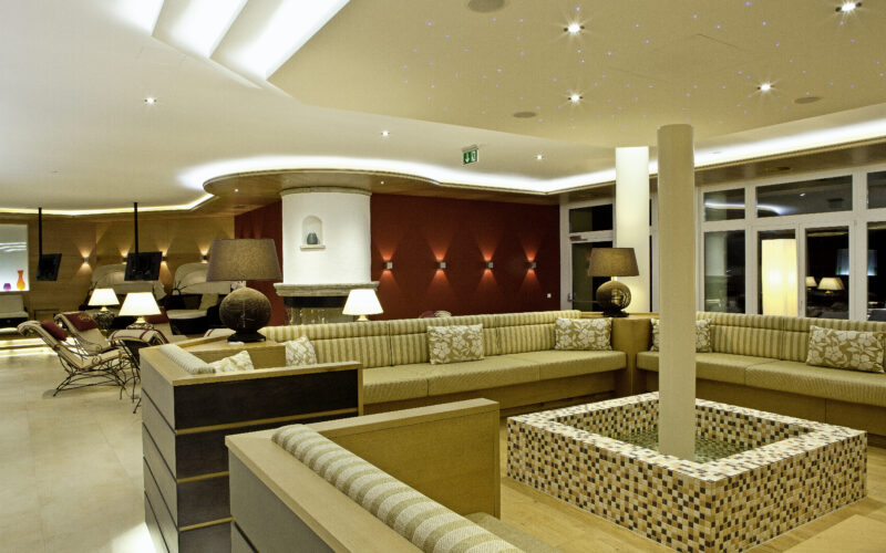 Moderne Hotellounge mit beigen Sektionssofas, Mosaikfliesenbrunnen, Ambientebeleuchtung und dekorativen Lampen. Die Decke ist mit sternförmigen Lichtern und einem geschwungenen Design versehen.