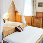 Ein gemütliches Hotelzimmer mit einem Doppelbett und einem Einzelbett, Holzmöbeln, dekorativen Teppichen, Vorhängen und natürlichem Licht durch große Fenster.