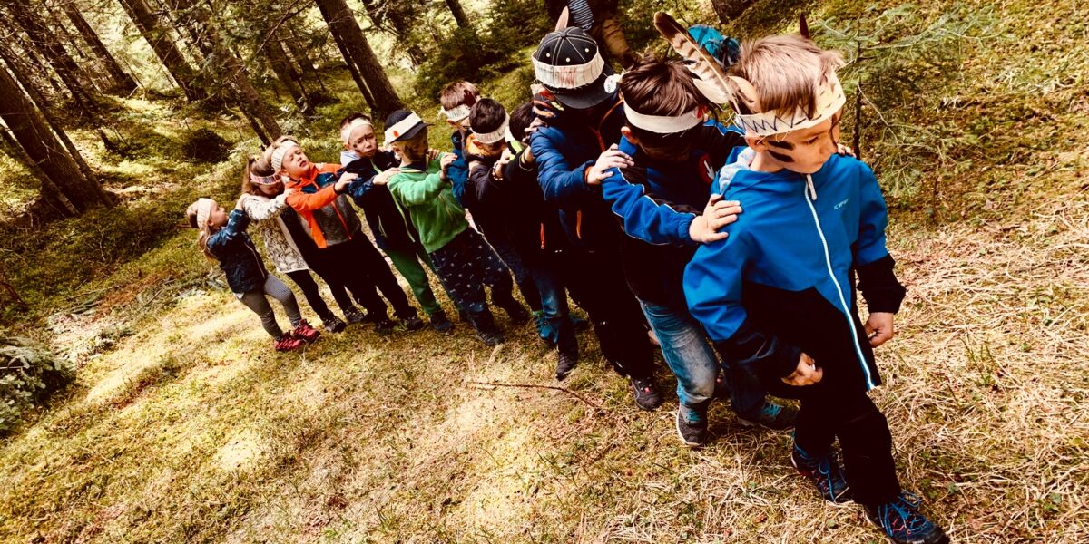 Eine Gruppe von Kindern, von denen einige die Augen verbunden haben, läuft in einer Reihe durch einen Wald, während ein Erwachsener im Hintergrund die Aufsicht führt.