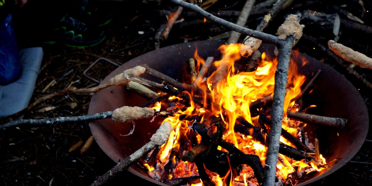 In einer metallenen Feuergrube brennt ein Lagerfeuer, in dem um Stöcke gewickelter Brotteig über den Flammen geröstet wird.