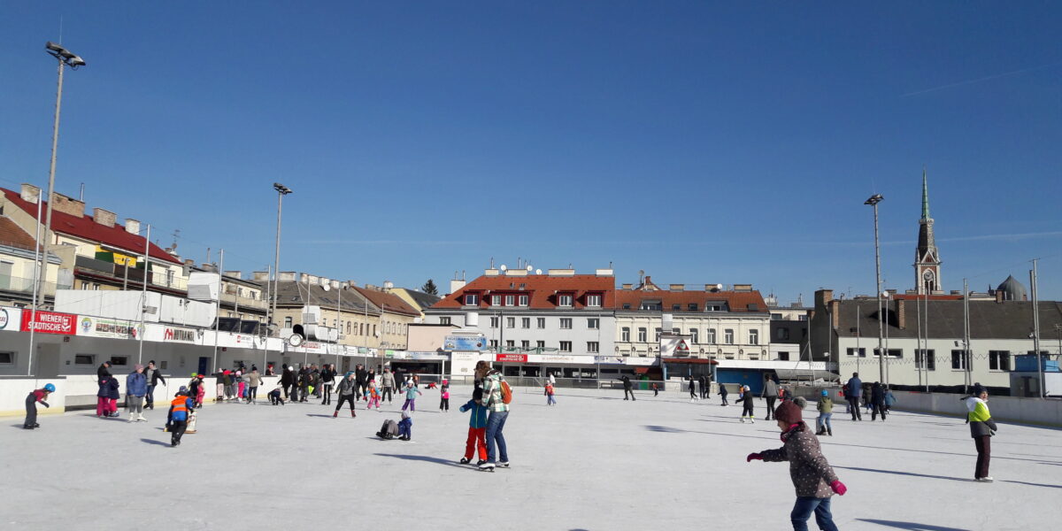 Menschen, die auf einer Eisbahn auf einem Stadtplatz Schlittschuh laufen, mit mehreren Gebäuden und einer Kirchturmspitze im Hintergrund unter einem klaren blauen Himmel.