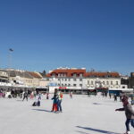 Menschen, die auf einer Eisbahn auf einem Stadtplatz Schlittschuh laufen, mit mehreren Gebäuden und einer Kirchturmspitze im Hintergrund unter einem klaren blauen Himmel.