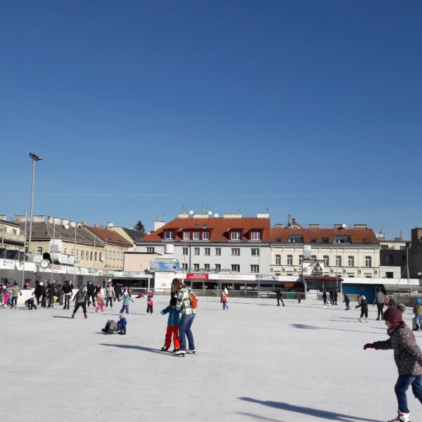 Menschen, die auf einer Eisbahn auf einem Stadtplatz Schlittschuh laufen, mit mehreren Gebäuden und einer Kirchturmspitze im Hintergrund unter einem klaren blauen Himmel.