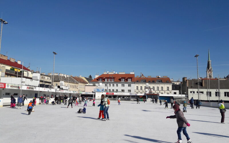 Menschen, die auf einer Eisbahn auf einem Stadtplatz Schlittschuh laufen, mit mehreren Gebäuden und einer Kirchturmspitze im Hintergrund unter einem klaren blauen Himmel.