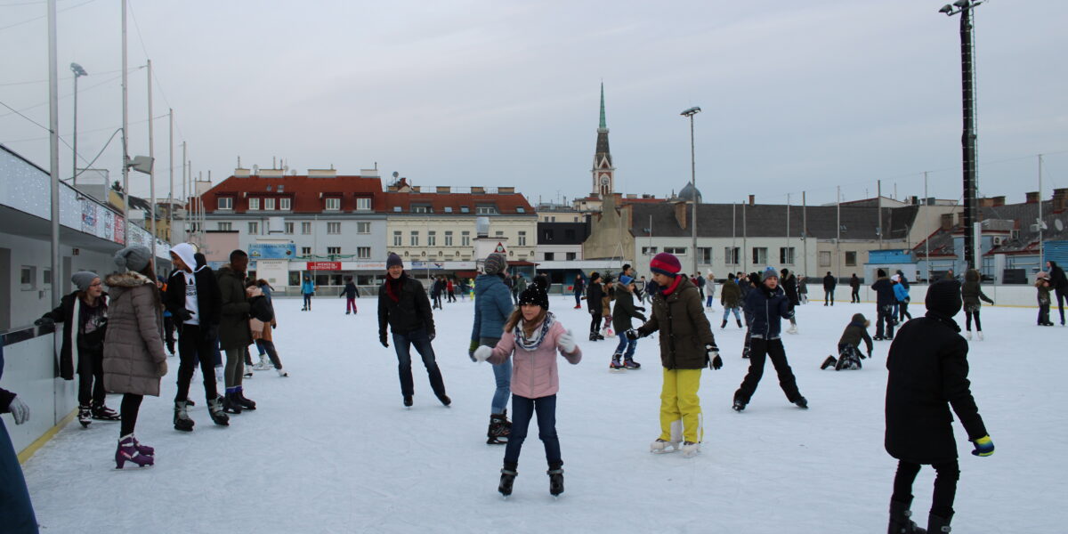 Menschen verschiedenen Alters beim Schlittschuhlaufen auf einer Freiluft-Eisbahn in einer Stadt, mit Gebäuden und einem Kirchturm im Hintergrund.