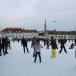 Menschen verschiedenen Alters beim Schlittschuhlaufen auf einer Freiluft-Eisbahn in einer Stadt, mit Gebäuden und einem Kirchturm im Hintergrund.